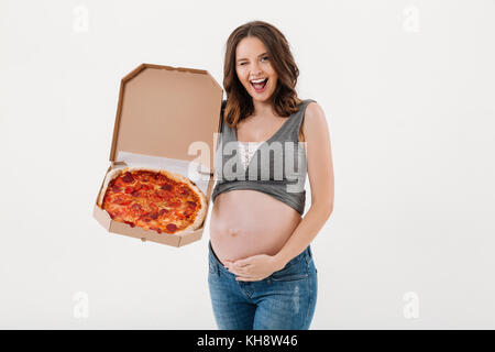 Image of happy pregnant woman standing isolated over white background. Looking camera holding pizza and winking. Stock Photo