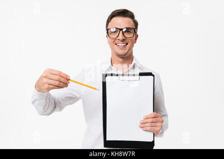 Portrait of a happy smart guy in white shirt and eyeglasses pointing with a pencil at blank clipboard and looking at camera isolated over white backgr Stock Photo