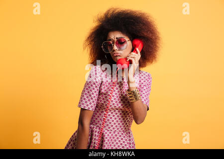 Close-up photo of upset retro girl with afro hairstyle posing with retro phone, isolated over yellow background Stock Photo