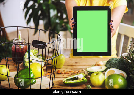 Woman showing a tablet with a green display at the kitchen table with fresh green vegetables and fruits, chroma copyspace Stock Photo