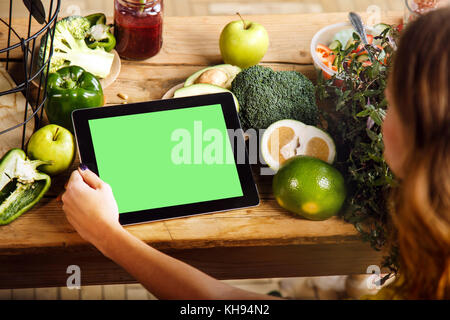 Woman showing a tablet with a green display at the kitchen table with fresh green vegetables and fruits, chroma copyspace Stock Photo