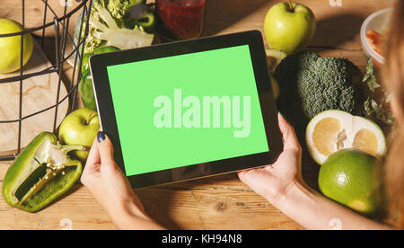 Woman showing a tablet with a green display at the kitchen table with fresh green vegetables and fruits, chroma copyspace Stock Photo
