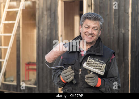 Senior man standing outside holding paint brushes and smiling. Stock Photo