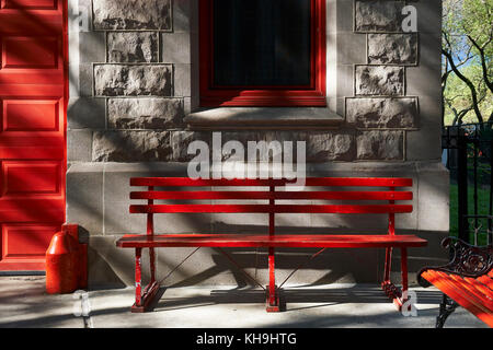 Evening light on red painted bench, under red window frame beside red door against gray stone wall Stock Photo