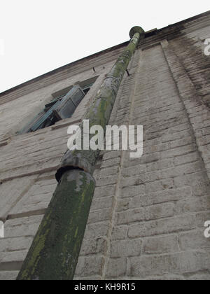 Green drainpipe, old, broken, with peeling paint on the stone wall of the house with a green wooden window, at the top empty space for text. Stock Photo