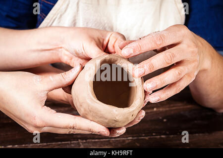 A close-up of a female potter teaches a woman in a denim shirt and apron molds a brown clay in the form of a ball, kneads the clay and makes a hole in Stock Photo