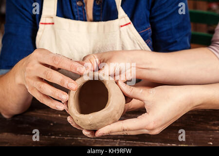 A close-up of a female potter teaches a woman in a denim shirt and apron molds a brown clay in the form of a ball, kneads the clay and makes a hole in Stock Photo