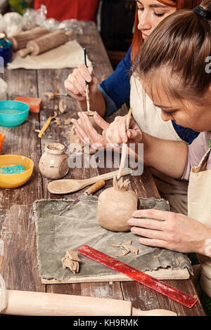 Pottery lesson: a woman potter paints a candlestick from clay, and another woman sculpts a candlestick on a wooden table with tools, brushes in a beau Stock Photo