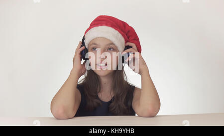 Beautiful naughty girl teenager in a Santa Claus hat listens to music on headphones on white background Stock Photo