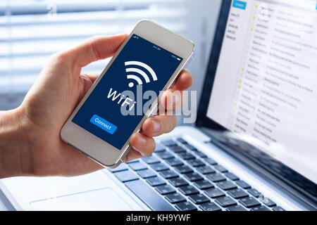 WiFi symbol on smartphone screen with button to connect to wireless internet, close-up of hand holding mobile phone, computer in background Stock Photo