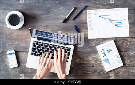 Project manager using laptop computer, printed Gantt chart planning with milestone and task schedule, smartphone and tablet computer with wireless int Stock Photo