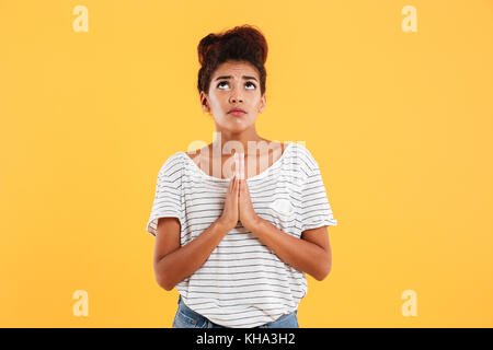 Young nervous pretty african lady looking up and praying while holding hands together isolated Stock Photo