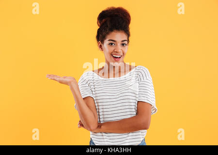 Happy african casual lady holding on her hand copy space and smiling to camera isolated over yellow Stock Photo