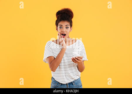 Pretty african surprised lady looking camera while using smartphone isolated over yellow Stock Photo