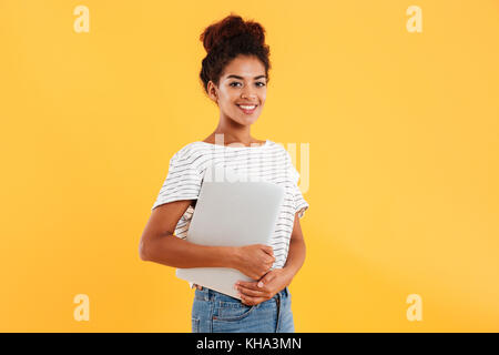 Pretty african lady holding laptop and smiling to camera isolated over yellow Stock Photo