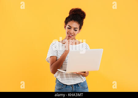 Young african positive cool lady with curly hair using laptop and smiling isolated over yellow Stock Photo