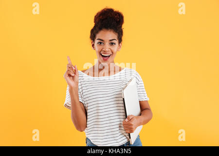 Pretty smiling african lady holding laptop and having an idea isolated over yellow Stock Photo