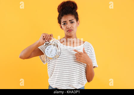 Upset african woman holding alarm clock and pointing on them over yellow background Stock Photo