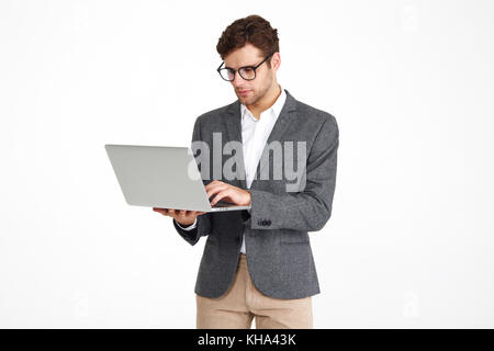 Portrait of a young concentrated businessman in eyeglasses and a jacket working on laptop computer while standing isolated over white background Stock Photo