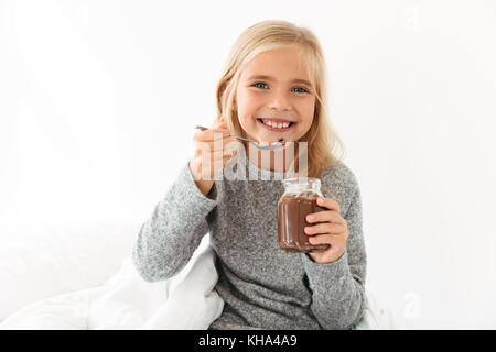 Happy blonde girl in gray pajamas holding glass jar and spoon with chocolate spread, looking at camera Stock Photo