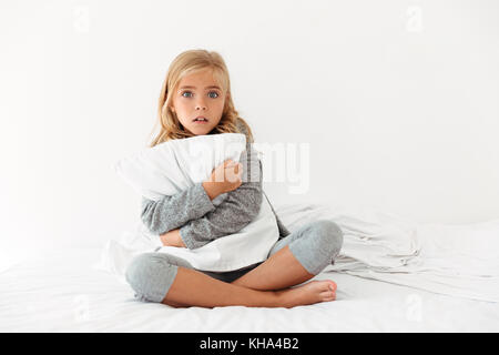 Portrait of a scared little girl hugging pillow while sitting on bed and looking at camera in the morning Stock Photo