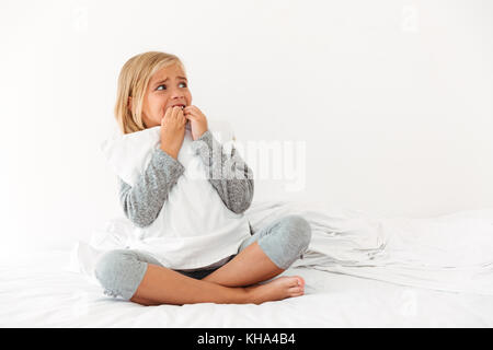 Portrait of a panicked little girl hugging pillow while sitting on bed and looking away in the morning Stock Photo