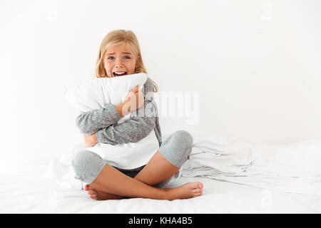 Portrait of a terrified little girl hugging pillow while sitting on bed and looking at camera in the morning Stock Photo