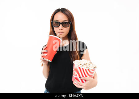 Portrait of a young asian girl in 3d glasses drinking from a cup with a straw while holding popcorn box and looking at camera isolated over white back Stock Photo