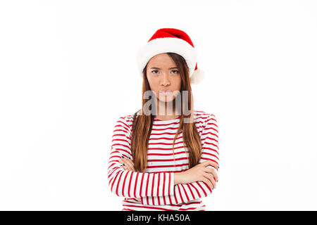 Portrait of an upset asian girl in christmas hat standing with arms folded isolated over white background Stock Photo