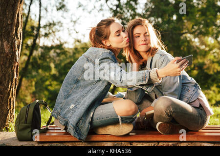 Two playful young woman in jeans wear making selfie while sitting on wooden bench in park Stock Photo