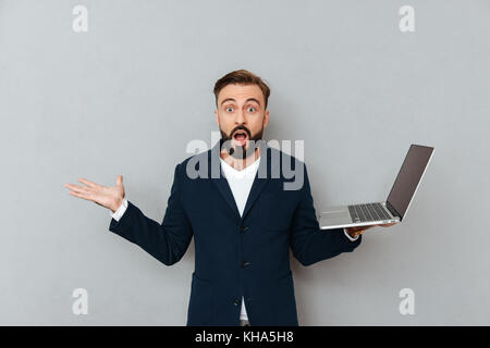 Shocked bearded man in suit holding laptop computer and looking camera with opened mouth isolated over grey Stock Photo