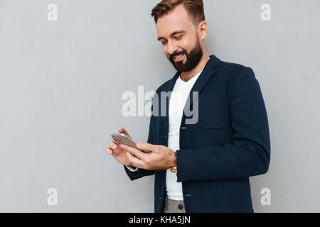 Smiling concentrated bearded man in suit using smartphone isolated over grey Stock Photo