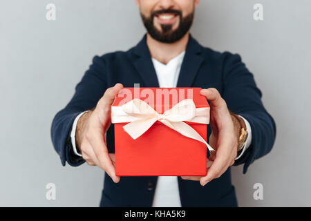 Cropped image of smiling bearded man in business clothes presents a gift at camera over gray background Stock Photo
