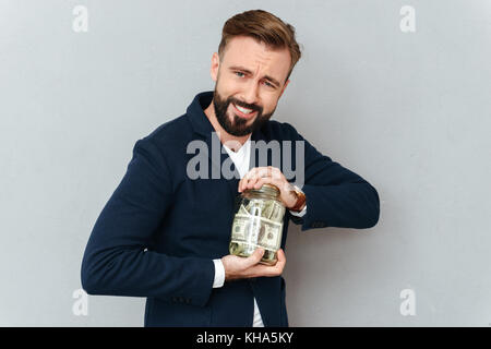 Concentrated smiling bearded man in business clothes trying opening jar with money over gray background Stock Photo