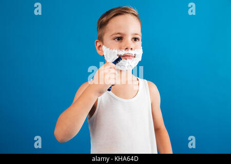 Young boy trying shaving his face like man and looking away over blue background Stock Photo