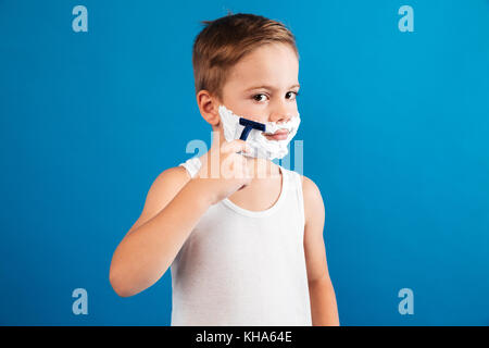 Young boy trying shaving his face like man and looking at the camera over blue background Stock Photo