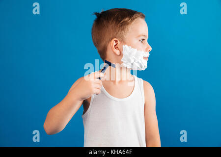 Young boy trying shaving his face like man and looking aside over blue background Stock Photo