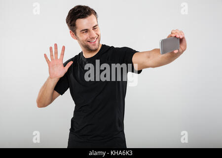 Portrait of a smiling young guy waving to camera holding mobile phone isolated over gray background Stock Photo