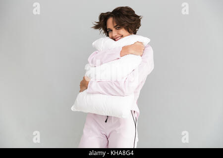 Portrait of a lovely young girl in pajamas hugging a pillow and looking at camera isolated over gray background Stock Photo