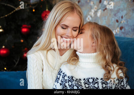Close-up photo of little girl in knitted sweater kiss her mother while sitting on sofa at Christmas tree Stock Photo