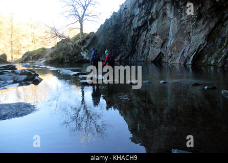 A Couple of Fell Walkers in Rydal Cave Walking on Stepping Stones above Rydal Water in the Lake District National Park, Cumbria, UK. Stock Photo