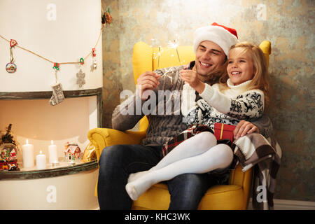 Cheerful little girl with her daddy holding sparklers, while sitting in chair near fireplace Stock Photo