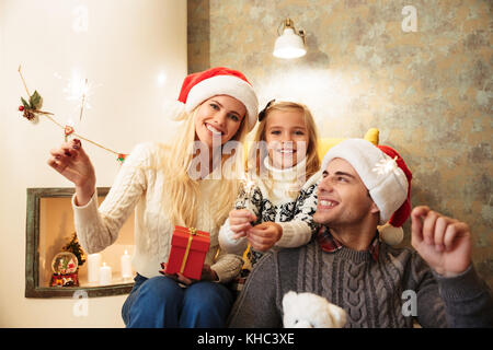 Happy beautiful famyly with daughter holding sparkler, looking at camera while celebrating Christmas Stock Photo