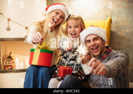 Smiling mother, father and little daughter holding sparklers, looking at camera while celebrating Christmas Stock Photo