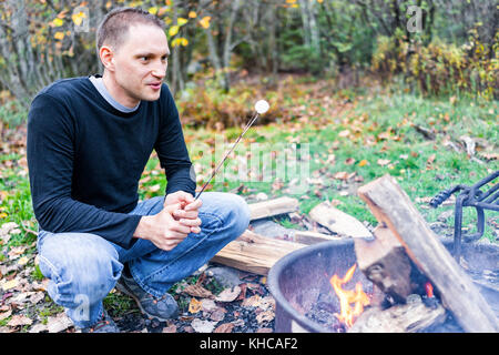 Closeup of young happy smiling man roasting one white marshmallow carmelizing on fire showing detail and texture by campground campfire grill in outdo Stock Photo