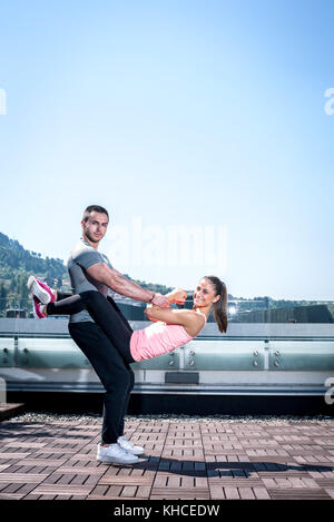 Young attractive couple doing advanced double dare crunches while standing on rooftop of the building in urban area. Stock Photo