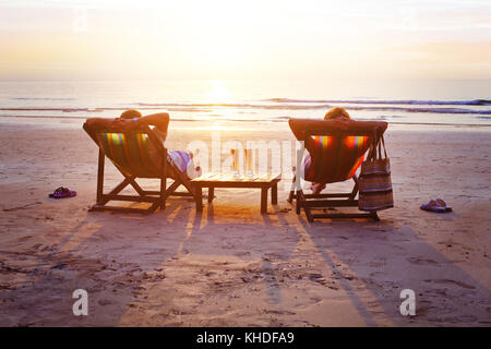 honeymoon travel, silhouettes of happy couple relaxing in deck chairs on the beach at sunset Stock Photo