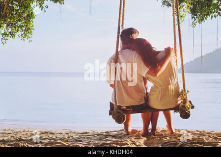romantic holidays for two, affectionate couple sitting together on the beach on swing, silhouette of man hugging woman Stock Photo