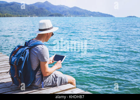 traveler using digital tablet computer while sitting on wooden pier, travel app online, man backpacker with wireless device Stock Photo