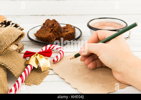 Woman hand writing a letter to Santa Claus on white table with winter holidays decorations, hot cholocate cup and brown cake Stock Photo
